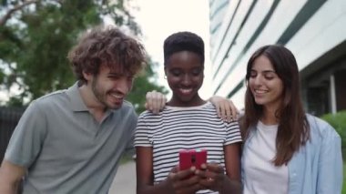 Young multiracial friends having fun looking at mobile phone screen outside. Diverse teenage people laughing together watching social media content on smart phone app while walking in city street.