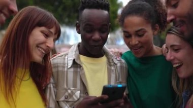 Young happy student friends having fun together using mobile phone standing outdoors. Multi-ethnic group of millennial people laughing while watching funny social media content on smartphone app.