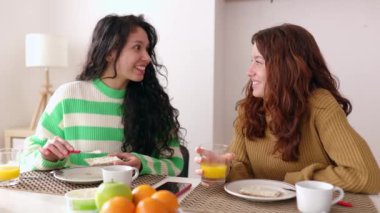 Young diverse female friends talking while having breakfast in the living room at home. Domestic lifestyle concept