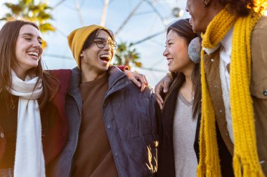 Happy young group of multiracial friends laughing while hugging each other outdoors. Millennial student people in winter clothes having fun together on vacation.