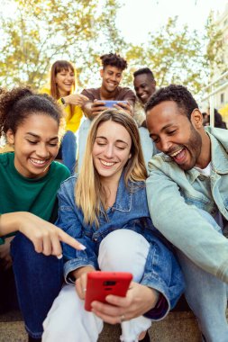 Vertical shot of united group of multiracial people watching funny content on mobile phone app. Millennial diverse friends having fun using smartphone devices outdoors