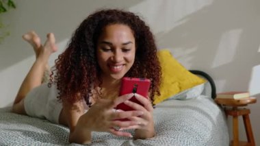 Smiling pretty young latin american woman browsing on mobile phone app. Happy teenage girl having fun using smartphone device while lying on bed in the morning at home.