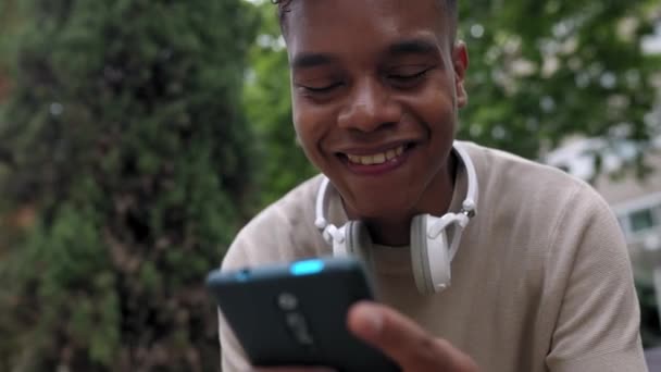 スマートフォンを使って楽しむアフリカ系アメリカ人の10代の少年のクローズアップ 都会の公園でスマートフォンを持つ面白いソーシャルメディアコンテンツを見ながら笑う楽しい千年の男 — ストック動画