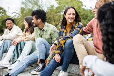Dışarıda otururken gülen çok ırklı bir grup birleşik genç arkadaş. Milenyum çeşitliliği olan insanlarla arkadaşlık öğrenci yaşam tarzı konsepti şehir sokaklarında eğlenceli sosyal buluşmalar yapıyor.
