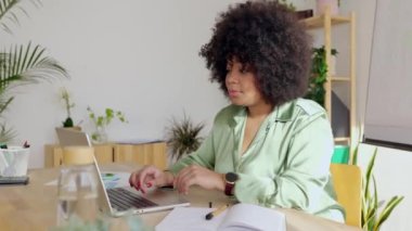 Young adult freelance Latin American black businesswoman taking notes while working on laptop computer sitting at bright modern business office.