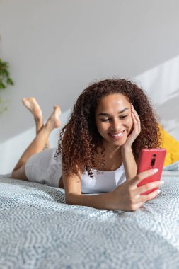 Vertical shot of smiling pretty young latin american woman browsing on mobile phone app. Happy teenage girl having fun using smartphone device while lying on bed in the morning at home.