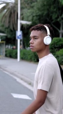 Yaya geçidinden geçen yayalar. Şehirde kulaklıkla müzik dinlerken bir zebranın üzerinde yürüyen genç ve kendinden emin bir Latin çocuğun dikey video görüntüsü..
