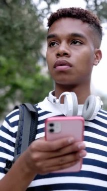 Vertical screen video of young hispanic latin man in striped shirt with headphones holding smart mobile phone standing outdoor looking for address with smartphone app in city street. HD shot footage