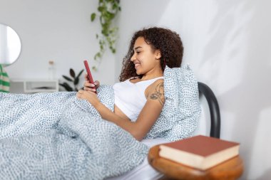 Side view of happy young latin american woman using mobile phone resting on bed in the morning. Technology and domestic lifestyle