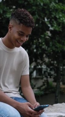 Vertical video shot of happy young hispanic latin teenager boy having fun chatting on smartphone app while sitting outdoor. Happy ethnic guy using cellphone to share media content.