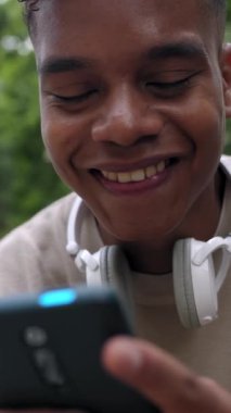 Genç Afro-Amerikan delikanlının akıllı telefon kullanarak eğlendiğini gösteren dikey video görüntüsü. Komik sosyal medya içeriğini izlerken gülen neşeli milenyum adamı parkta akıllı telefon tutuyor.