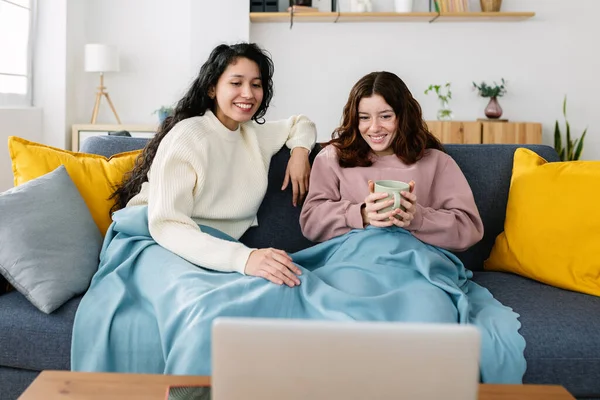 Two young female roommates covered with blue blanket relaxing on sofa watching movie on laptop. Friendship and multimedia streaming entertainment concept