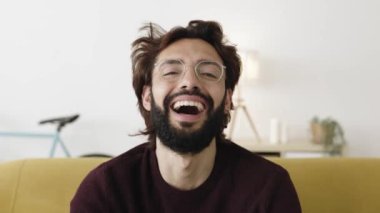 Evdeki kanepede otururken kameraya gülen mutlu sakallı bir gencin portresi. Mutluluk ve neşeli insanlar konsepti