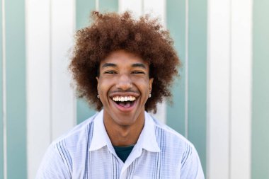 Cheerful young latino american man with curly hair in summer clothes laughing at camera outdoors clipart