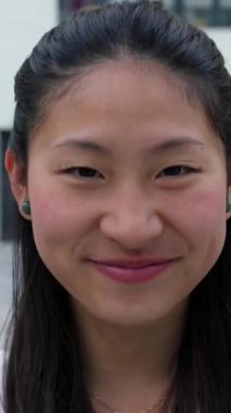 Şehir caddesinde yürürken kameraya bakıp gülümseyen Asyalı genç bir kadının dikey portresi. Neşeli insanlar konsepti