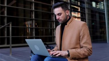 Laptopta çalışan yakışıklı genç sakallı iş adamı ofis binasının merdivenlerinde oturuyor..