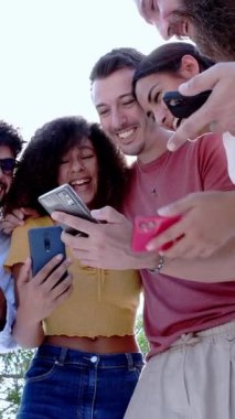 Birlikte cep telefonu kullanırken gülümseyen genç insanların dikey görüntüsü. Parkta elinde cep telefonuyla bekleyen çok ırklı arkadaşlar. Teknoloji, gençlik ve sosyal medya kavramı