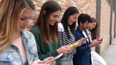 Tuğla duvara dayanan akıllı telefon kullanan çok ırklı bir grup genç arkadaş. Sosyal medya içeriğinden hoşlanan mutlu öğrenci çeşitliliği açık havada akıllı cep telefonu aygıtı. Teknoloji konsepti