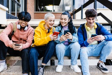 Dışarıdaki binanın merdivenlerinde cep telefonlarını kullanan mutlu, çok ırklı lise öğrencileri grubu. Teknoloji, sosyal medya ve halk konsepti.