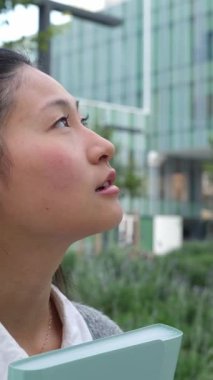 Asyalı genç bir kadının ofis binasının önündeki dikey videosu. Şehir sokağına bakıyor.. 