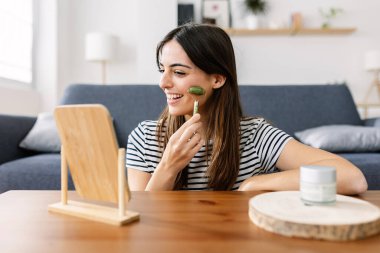 Gülümseyen mutlu genç bir kadın evde yüzüne masaj yapmak için yeşim kuvars yüz silindiri kullanıyor. Cilt bakımı kavramı