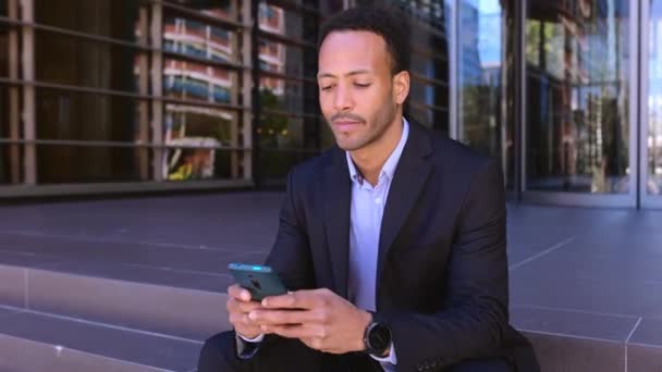 若い魅力的なアフリカ系アメリカ人のビジネスマン階段の建物の入り口に座って携帯電話を使用して — ストック動画