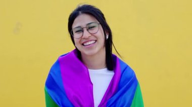 Sarı arkaplanda gülümseyen genç lezbiyen kadın portresi LGBTQ onur günü festivalini kutluyor. Eşcinsel olmaktan gurur duyan gökkuşağı bayrağıyla kaplı.