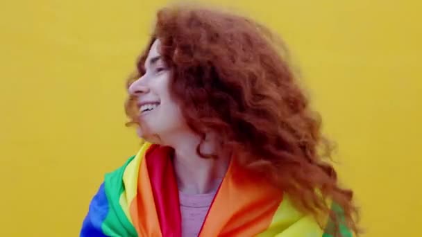黄色の背景にカメラで笑みを浮かべて虹のフラグを持つ赤い髪の非バイナリ男の肖像画 Lgbtコミュニティ多様性トランスジェンダーと非バイナリの人々の概念 — ストック動画