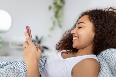 Side view of happy young latin american woman using mobile phone resting on bed in the morning. Technology and domestic lifestyle