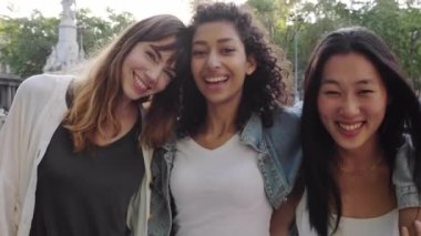 Modern şehrin arka planında birlikte yürürken kameraya gülümseyen üç genç ve çok ırklı kadın. Güzel, küçük bir bayan arkadaş grubu. Birbirlerine sarılıyorlar. Tatilin tadını çıkarıyorlar..