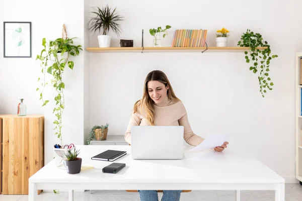 Laptop bilgisayarında çalışan genç bir kadın ofis masasında oturuyor. Milenyum sarışın iş kadını belgelere bakarken bilgisayar ekranına bakıyor. 