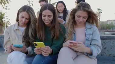 Dışarıda cep telefonu kullanan bir grup mutlu genç öğrenci. Çok ırklı insanlar cep telefonlarındaki ağ uygulama platformunda sosyal medya içeriğini izlerken eğleniyor.