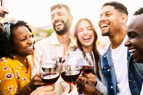 Multiracial group of young friends toasting red wine enjoying summer party at terrace house. Happy people having fun together on birthday patio celebration. Friendship lifestyle concept.