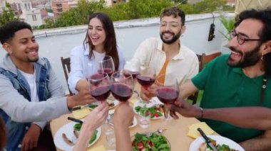 Bir grup gülümseyen insan teras partisinde kırmızı şarap kadehleriyle kadeh kaldırıyor. Mutlu arkadaşlar gülüyor ve eğleniyor. Yazın çatıda öğle yemeği kutlaması yapıyorlar..