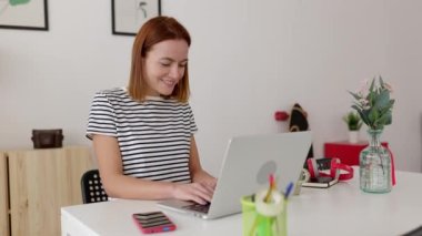 Oturma odasındaki masada oturan neşeli genç kadın dizüstü bilgisayarla çalışıyor. Milenyum kız öğrencisi evden online olarak çalışıyor. Teknoloji yaşam tarzı ve eğitim kavramı.