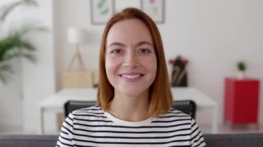 Genç ve güzel bir kadın kanepeye oturmuş laptoptan video dinliyormuş. Vesikalık kamera görüntüsü. Sanal toplantı konferansına katılan gülümseyen kadın portresi..