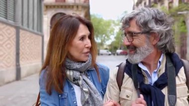 Yaşlı evli bir çiftin şehirde yürüyüp konuşmalarını kameraya gülümsemelerini ön planda görmek. Tatilde eski kasabayı gezerken mutlu yaşlı turistler.