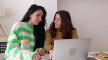 Gülümseyen genç yetişkin milenyum kız arkadaşları evdeki oturma odasındaki masada oturan dizüstü bilgisayarla birlikte öğreniyorlar. Eğitim ve arkadaşlık kavramı
