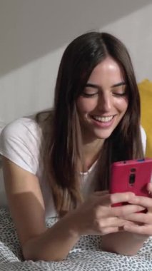 Dikey video. Sabahları yatağında cep telefonu kullanan genç bir kadın. Gülümseyen güzel bir milenyum kadını odada dinlenirken akıllı telefondan sohbet ediyor. Teknoloji ve ev hayatı kavramı