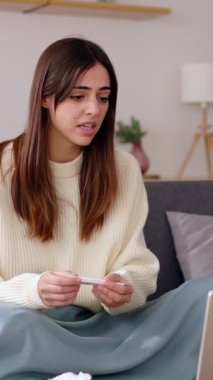 Ateşini ölçen, alnına dokunan genç hasta bir kadın evdeki oturma odasındaki kanepede oturan doktorla görüntülü konuşma yapıyor. Dikey video.