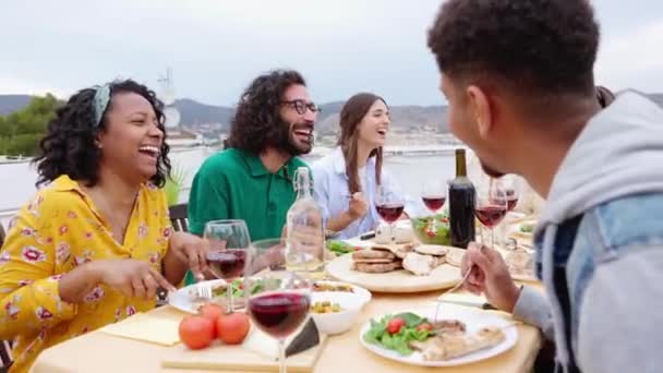 夏のパーティーのお祝いで一緒に食事をしている友人の幸せな若いグループ 屋上テラスでバーベキューや赤ワインを楽しむ多人種男性と女性 — ストック動画