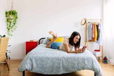 Yatakta cep telefonu kullanan mutlu genç bir kadın. Rahatlamış güzel milenyum dişisi cep telefonuyla sohbet ediyor evde dinleniyor.