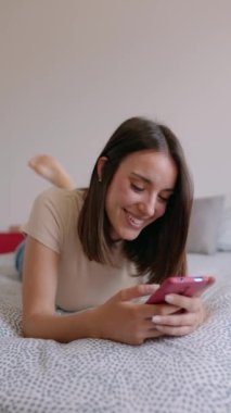 Evde dinlenirken cep telefonu kullanan güzel bir kadının dikey portresi. Yatakta uzanmış akıllı telefon ekranına bakarak gülümseyen güzel kız. Sosyal medya ve teknoloji kavramı.