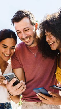 Birlikte cep telefonu kullanırken gülümseyen bir grup mutlu insan. Parkta elinde akıllı telefon cihazlarıyla bekleyen çok ırklı gülen arkadaşlar. Teknoloji, gençlik ve sosyal medya kavramı