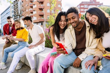 Çok ırklı bir grup genç, açık havada cep telefonu kullanırken eğleniyor. Gülümseyen öğrenci topluluğu ellerinde akıllı cep telefonları olan insanlar şehrin caddesinde birlikte oturuyorlar..