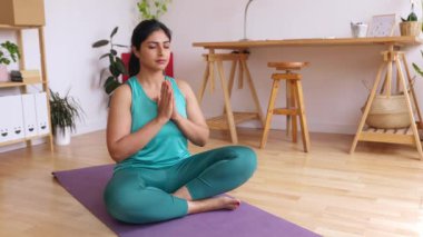 Huzurlu Hintli etnik kadın yoga minderinde bağdaş kurarak oturuyor. Ev stüdyosunda gözleri kapalı nefes alıp meditasyon yapıyor..