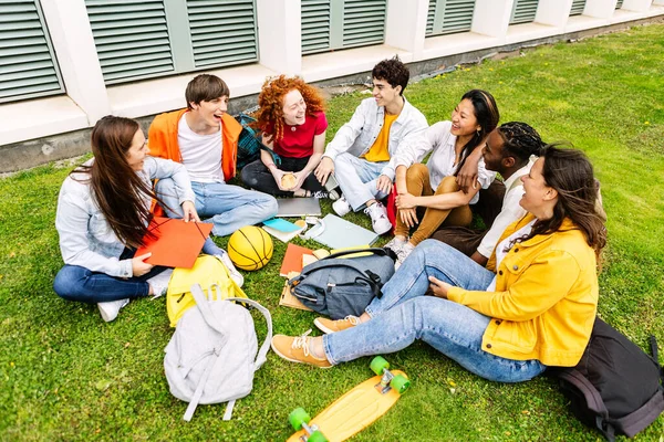 Happy student college friends laughing and relaxing together sitting on campus grass. Multiracial group of classmate people social gathering after university classes. Education and youth concept.