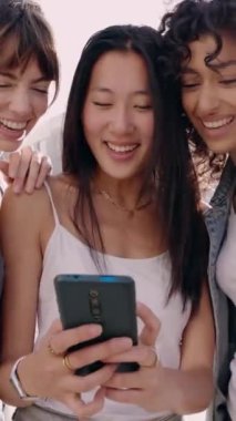 Cep telefonunda sosyal medya uygulaması kullanarak eğlenen üç etnik kadın. Küçük bir grup bayan arkadaş şehrin arka planındaki akıllı telefon ekranına bakarken gülümsüyor. Dikey video.