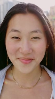 Şehirli genç Asyalı kızın portresi kameraya gülümsüyor. Açık havada gülen neşeli Çinli kadının yakın çekimde görüntüsü. Dikey video görüntüleri.