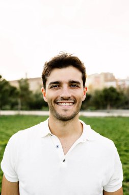 Şehir parkında kameraya bakan yakışıklı bir gencin dikey portresi. Dışarıda dikilen mutlu milenyum adamı.